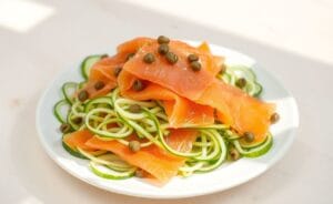 Salada de pepino em fatias finas com salmão defumado, alcaparras e um leve molho, servida em prato branco.