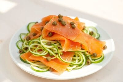 Salada de pepino em fatias finas com salmão defumado, alcaparras e um leve molho, servida em prato branco.